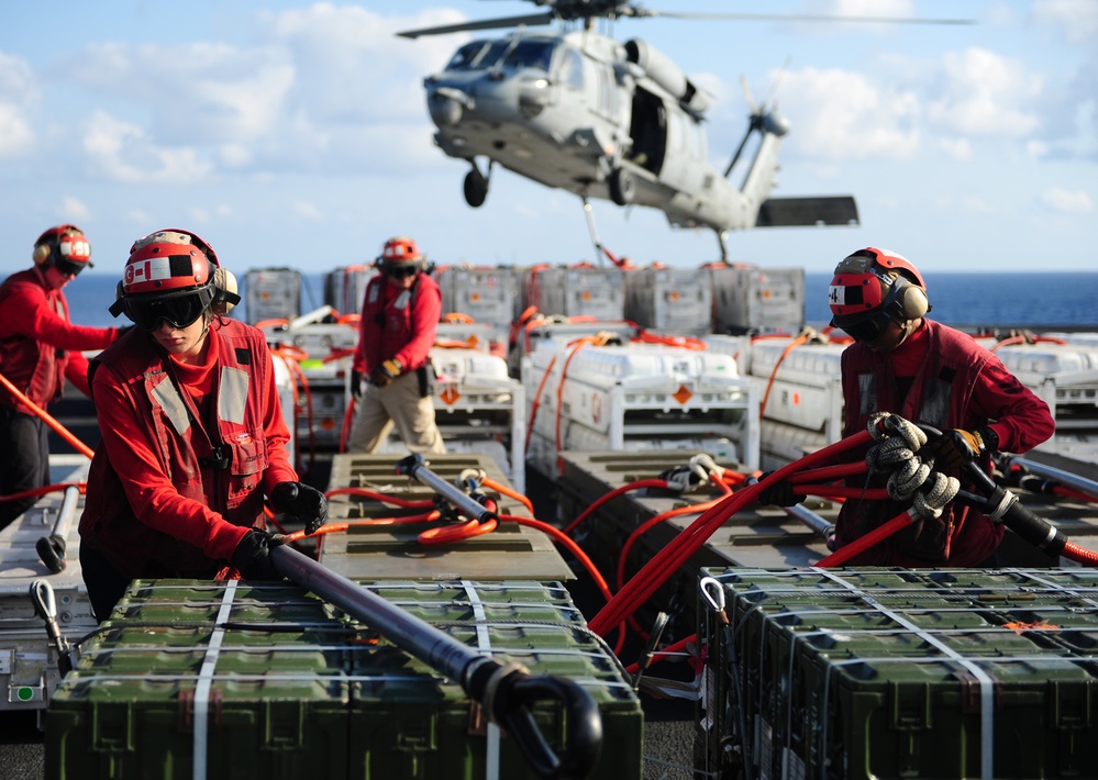 Military personnel in red uniforms and safety helmets are conducting a replenishment-at-sea (RAS) or vertical replenishment (VERTREP) operation on the deck of a naval vessel. Crew members are handling red hoses and securing cargo containers while a military helicopter hovers overhead. The scene depicts a coordinated logistics operation transferring supplies between vessels or from helicopter to ship.
