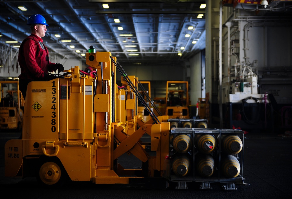 A worker wearing a blue hard hat and red uniform operates a yellow industrial forklift inside what appears to be the hangar bay of a naval aircraft carrier. The forklift, marked with the number '1324380' and labeled 'Shipboard Use Approved', is carrying a rack of cylindrical objects, likely ordnance or pressurized canisters. The large, well-lit interior space contains multiple additional yellow forklifts and industrial equipment in the background.