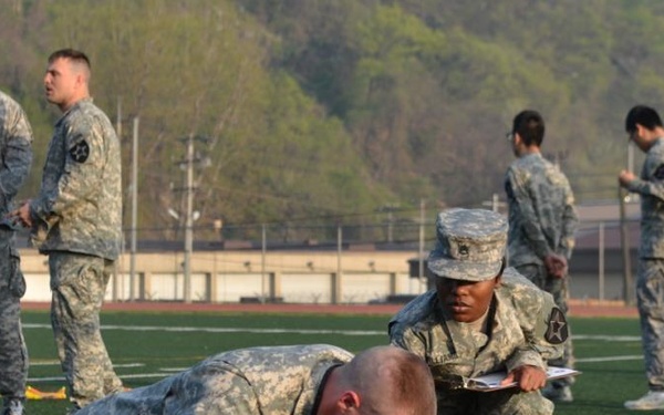 A group of U.S. Army soldiers are conducting a physical fitness test on an outdoor athletic field. One soldier wearing number '32' is performing push-ups while another soldier in a patrol cap observes and records the count on a clipboard. Additional soldiers are visible in the background, some appearing to supervise or await their turn.