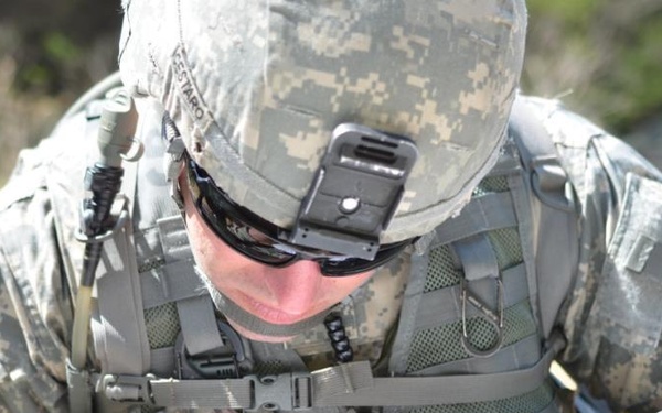 A U.S. Army soldier in ACU (Army Combat Uniform) digital camouflage is bent over a topographic map, performing land navigation. The soldier is wearing a combat helmet with a mounted device, tactical vest, and sunglasses. Visible unit patches identify affiliation with the 2nd Infantry Division and what appears to be the 42nd Infantry Division or similar unit.