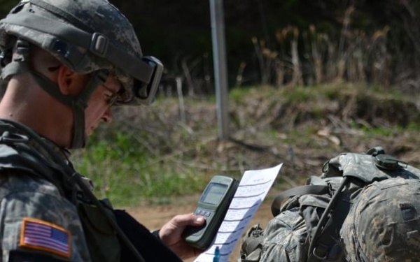 Two U.S. Army soldiers in ACU (Army Combat Uniform) camouflage are engaged in what appears to be a land navigation or field training exercise. Soldier number 32 is holding a handheld GPS device and a printed document, while soldier number 41 is bent over, likely writing or consulting a map. A third soldier is visible in the background also reviewing documents.