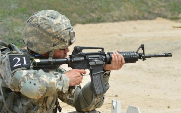 A U.S. Army soldier wearing ACU (Army Combat Uniform) and full tactical gear, numbered '21', is in a kneeling firing position on a military shooting range. The soldier is aiming an M16A2/M4-style rifle downrange, with a spent brass casing visible in the air to the upper right, indicating active firing. Lane marker '1' is visible in the foreground, confirming a structured range qualification or competition event.