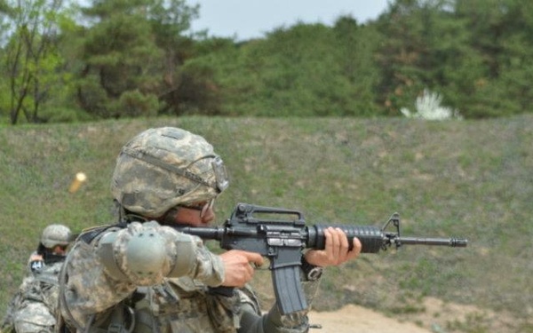 Two soldiers in U.S. Army Universal Camouflage Pattern (UCP) uniforms are engaged in a live-fire range exercise. The foreground soldier is actively firing an M16/M4-style rifle, with a visible muzzle flash or ejected casing near the weapon. The setting appears to be a military training range with earthen berms and a sandy firing lane.