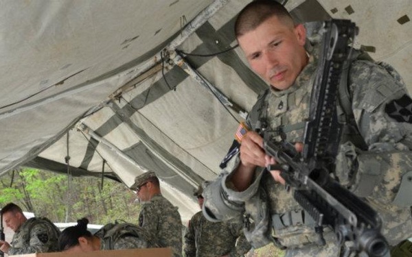 A group of U.S. Army soldiers are gathered under a military field tent at what appears to be a weapons maintenance or inspection station. The foreground soldier, wearing ACU (Army Combat Uniform) with a 2nd Infantry Division patch, is inspecting or handling an automatic weapon, likely an M249 SAW or similar light machine gun. Several weapons and cardboard boxes are laid out on a folding table, suggesting an armory check-in or pre/post-range activity.