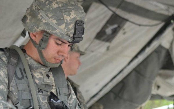 A U.S. Army soldier in ACU (Army Combat Uniform) is inspecting or handling an M4 carbine rifle under a field tent or canopy. The soldier wears a combat helmet with a mounted device and a name tape partially visible reading 'DUNCAN'. Additional soldiers in similar uniforms are visible in the background, suggesting a training or qualification range event.