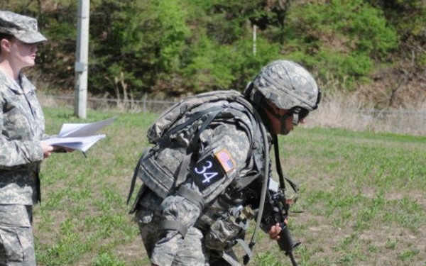A military training exercise is taking place in an open field. A soldier numbered '34' in full combat gear is practicing a casualty drag technique on a training mannequin dressed in military uniform. A second soldier, acting as an evaluator or observer, stands nearby holding a clipboard or papers.