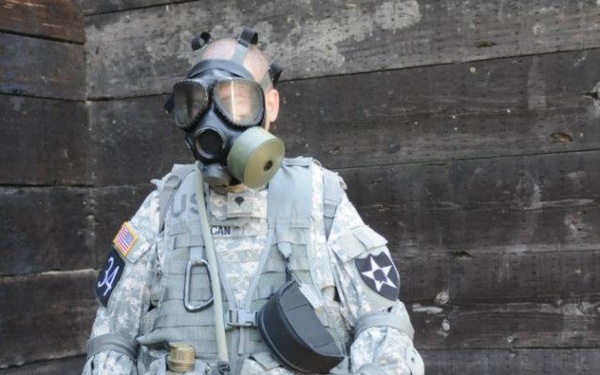 A U.S. Army soldier stands in full combat gear including a gas mask (M40 or M50 series), Army Combat Uniform (ACU) in Universal Camouflage Pattern, and a tactical vest with multiple pouches. The soldier is wearing the 2nd Infantry Division patch on the right shoulder and an American flag patch on the left. A rifle (M4 carbine) and a gray rucksack are visible on the ground beside them.