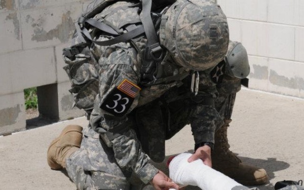 A soldier wearing US Army ACU (Army Combat Uniform) with the number '33' on their back is administering first aid to a simulated casualty lying on the ground. The standing soldier is applying a white bandage/dressing to the casualty's arm, with medical supplies scattered nearby. This appears to be a combat lifesaver or tactical combat casualty care (TCCC) training exercise.