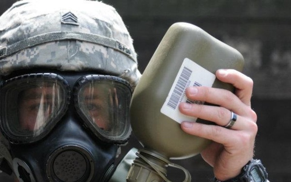 A U.S. Army soldier wearing an ACU (Army Combat Uniform) and an M40-series gas mask holds up an olive-drab military canteen or container toward the light. The soldier's rank insignia (Sergeant chevrons) and partial name tape ('MUTH') are visible on the uniform. The image appears to document CBRN (Chemical, Biological, Radiological, Nuclear) training or equipment inspection.