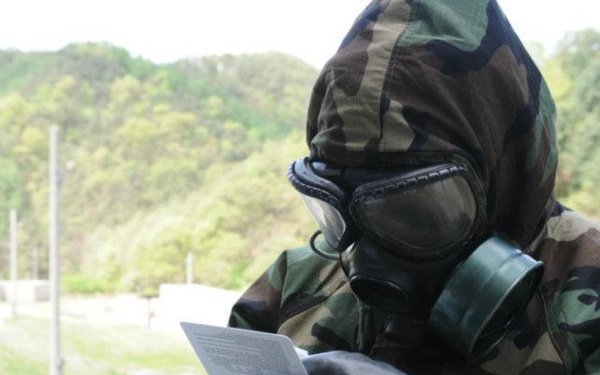 A person wearing full CBRN (Chemical, Biological, Radiological, Nuclear) protective gear, including a military gas mask and woodland camouflage suit, is examining what appears to be a chemical detection test strip against a color reference card. The individual is wearing black protective gloves and holds a white test strip in one hand and a color comparison chart in the other. The setting appears to be an outdoor military training or exercise environment.