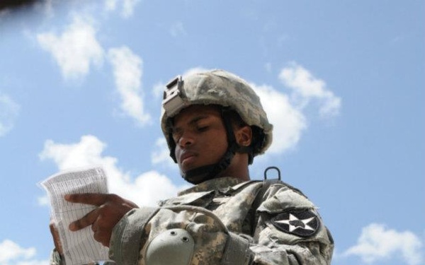 A U.S. Army soldier in ACU (Army Combat Uniform) is performing what appears to be a medical training exercise, attending to a mannequin or simulated casualty. The soldier is wearing a combat helmet and body armor with a unit patch visible on the left shoulder, and is reading from a document or card. The setting appears to be an outdoor training environment with a concrete structure in the background.