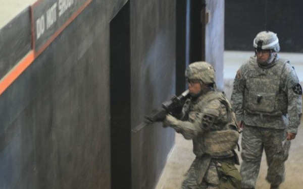 Two soldiers in US Army ACU (Army Combat Uniform) are conducting what appears to be a military training exercise in an indoor facility. The forward soldier is in a tactical crouch position holding a rifle, approaching a wall corner, while the second soldier stands upright observing. The setting appears to be a mock urban training environment with sandy flooring and constructed walls.