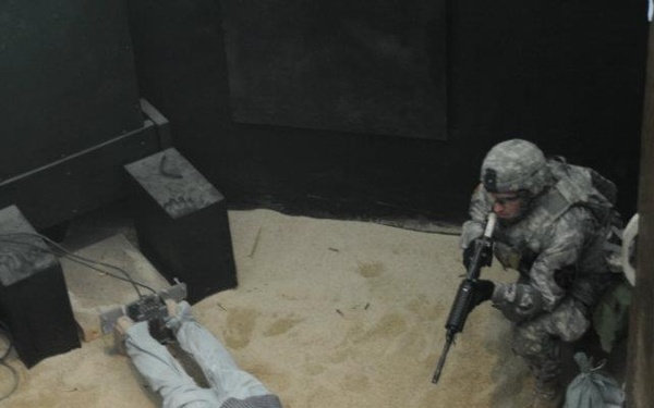 This appears to be a military training exercise or diorama/display scene set in a sand-floored room. A soldier in US Army ACU (Army Combat Uniform) is in a crouched tactical position holding a rifle, while a mannequin or training dummy in civilian clothes lies prone on the ground with hands bound. The setting includes props such as crates, sandbags, and wiring consistent with a training scenario.