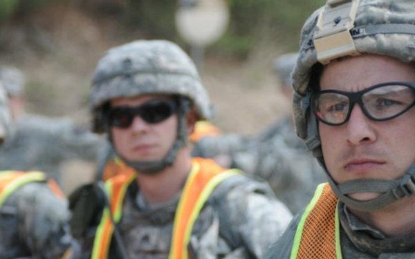 The image shows a group of U.S. Army soldiers in ACU (Army Combat Uniform) wearing orange high-visibility safety vests over their gear, suggesting a training exercise or safety detail. The foreground soldier is identified by a name tape on his vest as 'SSG Richardson' and wears ballistic glasses and a combat helmet. Multiple soldiers are visible in formation, with at least two carrying rifles.