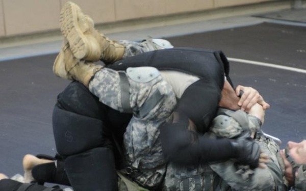 Two individuals are engaged in a ground grappling/combatives training session on a blue wrestling mat. One person is wearing US Army ACU (Army Combat Uniform) with a helmet and knee pads, while the other is dressed in a black training suit. The scene appears to be a military combatives training exercise conducted indoors.
