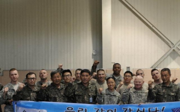 A group of approximately 20 military personnel from the United States and South Korea pose together for a group photo inside what appears to be a military facility or gymnasium. They are holding a banner that reads 'We Go Together' in English and '우리, 같이 갑시다!' in Korean, with both the South Korean Taegukgi and American flags displayed on the banner. The image represents a joint military cooperation or combined exercise event between US and ROK (Republic of Korea) forces.