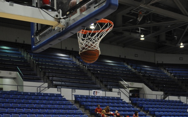 A wheelchair basketball game or practice session is taking place in an indoor arena, likely at the Warrior Games based on visible signage. A player in black is shooting a basketball toward the hoop, with the ball visible just below the rim. The arena seats are largely empty, suggesting this is a practice or warm-up session.