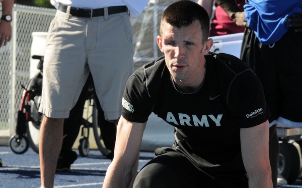 An athlete wearing a black Army-branded Nike shirt is positioned in a sprinter's starting stance on a blue athletic track, with feet placed on red starting blocks. The setting appears to be a competitive track and field event, likely a military or veterans' athletic competition. Other participants, officials, and equipment including a wheelchair are visible in the background.