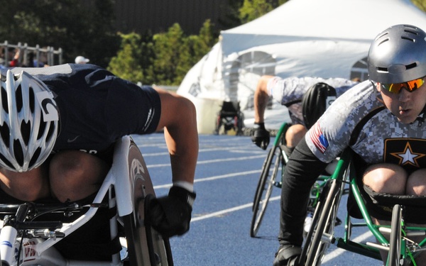 Two athletes are competing in a wheelchair racing event on a blue athletic track. The foreground racer wears a white aerodynamic helmet and dark jersey, while the second racer wears a gray digital camouflage jersey with a U.S. Army star logo and an American flag patch, suggesting a military adaptive sports competition. Both athletes are in aggressive forward-leaning racing positions using specialized racing wheelchairs.