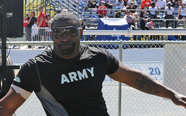 An Army athlete competes in a discus throwing event at what appears to be an adaptive sports competition, likely the Warrior Games or a similar military adaptive athletics event. The athlete is seated in a throwing chair on a grass field, mid-throw with a yellow discus in hand. A stadium with spectators is visible in the background.