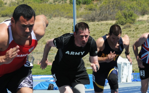 Four athletes are exploding off starting blocks on a blue track in what appears to be a Paralympic or military adaptive sports competition. The competitors represent different military branches and nations, including USMC, U.S. Army, and Great Britain. Several athletes are visibly using prosthetic limbs, highlighting this as an adaptive athletics event.