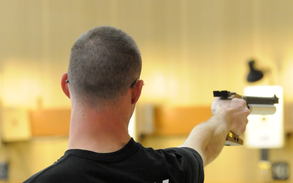 A competitor wearing an 'ARMY' black t-shirt participates in a pistol shooting event at the Warrior Games, sponsored by Deloitte. The athlete, wearing bib number 163 representing the USA, is shown from behind in a one-handed shooting stance aiming downrange at targets. The setting appears to be an indoor shooting range.
