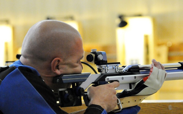 A bald male shooter is in a prone or bench-rested position at an indoor shooting range, aiming a precision target rifle downrange. He is wearing a specialized shooting jacket (white and blue) and a shooting glove, indicating competitive marksmanship. The background shows multiple illuminated target bays typical of an indoor rifle range.