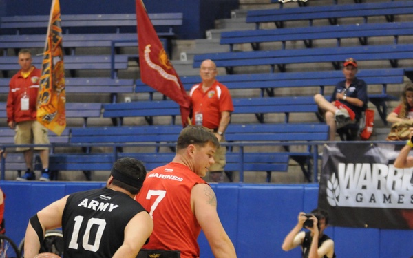 A wheelchair basketball game is taking place at the Warrior Games, featuring players from the Army and Marines competing on an indoor court. Two players are prominently visible in the foreground — an Army player (#10 in black) handling the ball and a Marines player (#7 in red) defending. Spectators, photographers, military flags, and a 'Warrior Games' banner are visible in the background.