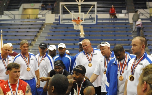 A wheelchair basketball ceremony or post-game gathering featuring military personnel from the Army team in black uniforms. Multiple participants are wearing medals with red, white, and blue ribbons, suggesting a competition awards ceremony. The event appears to be a Warrior Games or similar adaptive sports competition held in an indoor arena.
