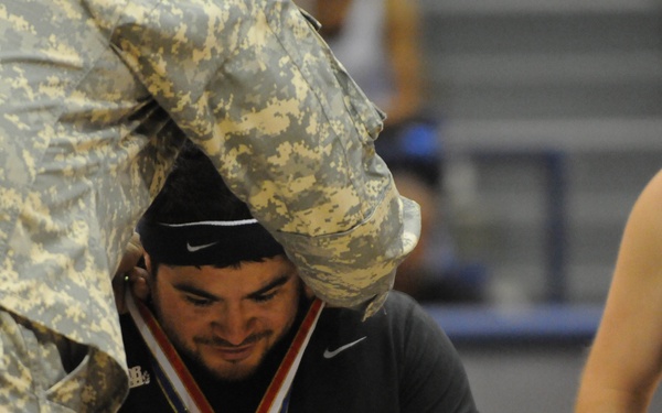 A medal ceremony is taking place, likely at a Warrior Games or similar adaptive sports competition. A person in military camouflage uniform is placing a gold medal around the neck of an athlete wearing a black Nike headband and a black jersey with a Deloitte sponsor patch. A second athlete wearing a black jersey numbered '5' with 'WAR GA' (likely Warrior Games) text is visible in the background.