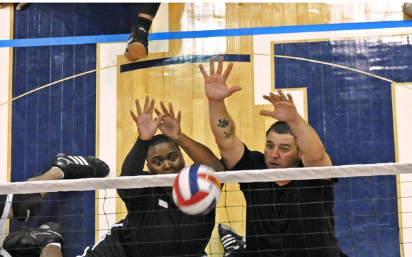 An overhead action shot of a sitting volleyball match, showing players competing on a gymnasium floor. A player in a red jersey (bib number 221) is spiking the ball toward the net, while two opposing players in black jerseys attempt a block. The image captures an intense moment of athletic competition, consistent with a Paralympic or adaptive sports event.