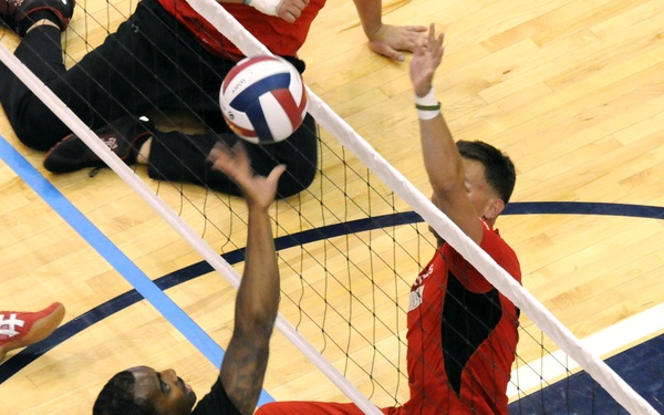 Two athletes are competing in a sitting volleyball match, a Paralympic sport played on the floor. A player in a black 'ARMY' jersey is spiking the ball over the net while an opponent in a red uniform attempts to block. Additional players are visible in the background seated on the court.