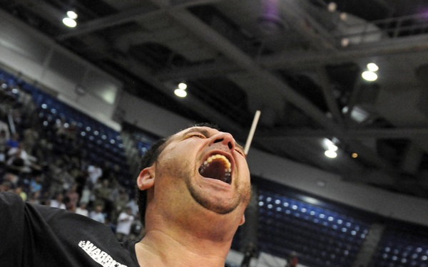 A man wearing a black Nike t-shirt with 'WARRIOR' text is captured mid-celebration, mouth wide open and head tilted back in an expression of intense joy or triumph. The setting is an indoor sports arena with a hardwood court floor, visible seating, and overhead lighting. Other participants in red shirts are visible in the background on the court.