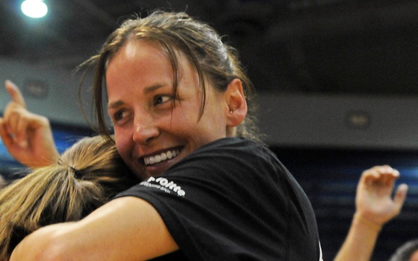 A group of people wearing matching black t-shirts are celebrating enthusiastically in what appears to be an indoor arena or gymnasium. The central focus is two women embracing in a joyful hug, with one woman smiling broadly at the camera. Two men in the background are also visibly excited, one pointing upward and the other smiling widely.
