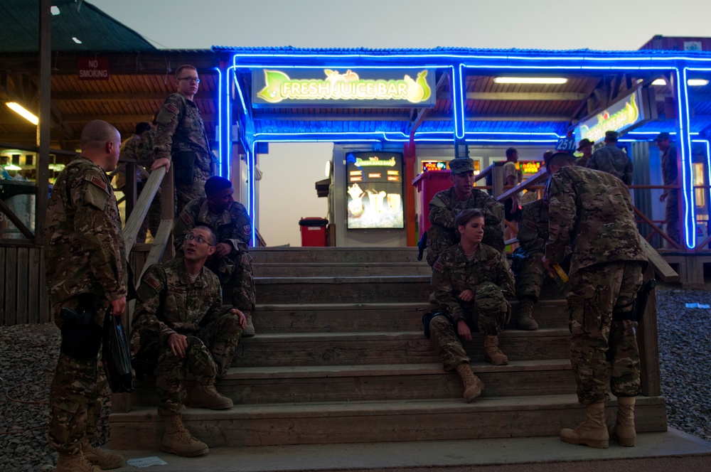 A group of military personnel in camouflage uniforms are gathered on wooden steps outside a 'Fresh Juice Bar' illuminated with blue neon lighting, likely on a forward operating base or military installation. The scene appears to be taken at dusk or early evening, with soldiers sitting, standing, and socializing in a recreational area. The setting suggests a designated rest and recreation zone within a military compound.