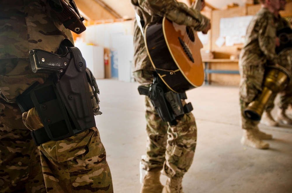 The image shows military personnel in camouflage uniforms inside what appears to be a forward operating base or military facility. One soldier is playing an acoustic guitar while others stand nearby, all wearing tactical gear including thigh holsters with sidearms. The scene depicts an informal moment of recreation or morale-boosting activity among deployed troops.