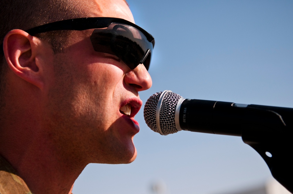 A close-up profile shot of a man singing or speaking into a professional microphone outdoors. He is wearing dark wraparound sunglasses and appears to be performing under bright sunlight with a clear blue sky in the background. The image is tightly cropped, focusing on his face and the microphone.