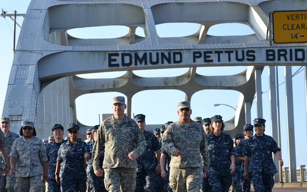 A group of U.S. military personnel in various service uniforms are marching across the historic Edmund Pettus Bridge in Selma, Alabama. The group includes members wearing Army Combat Uniforms (ACU), Air Force Airman Battle Uniforms (ABU), and Navy Working Uniforms (NWU). This appears to be a commemorative or ceremonial march across the bridge, which is historically significant as the site of the 1965 Selma to Montgomery civil rights marches.