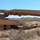 A military armored vehicle, identified as an M60 AVLB (Armored Vehicle Launched Bridge) or similar bridgelayer variant, is deploying a scissor-type bridge in an arid desert training environment. A soldier in combat uniform stands nearby observing the operation. The setting appears to be a military training range based on the terrain and conditions.