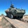 A U.S. Army Stryker wheeled armored fighting vehicle is crossing a metal bridge or ramp structure in an arid desert training environment. The vehicle bears unit markings and is equipped with a remote weapon station or turret-mounted weapon system. At least one soldier is visible in the turret, and another figure can be seen in the background on the left.