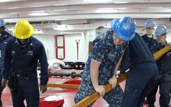 U.S. Navy sailors are working in what appears to be a ship's chain locker or anchor handling area, identifiable by the large anchor chain links in the foreground. Personnel wearing blue camouflage uniforms and hard hats of varying colors are using a wooden bar or timber to manipulate or guide heavy anchor chain on a red non-slip deck surface. The setting is an interior compartment of a naval vessel.