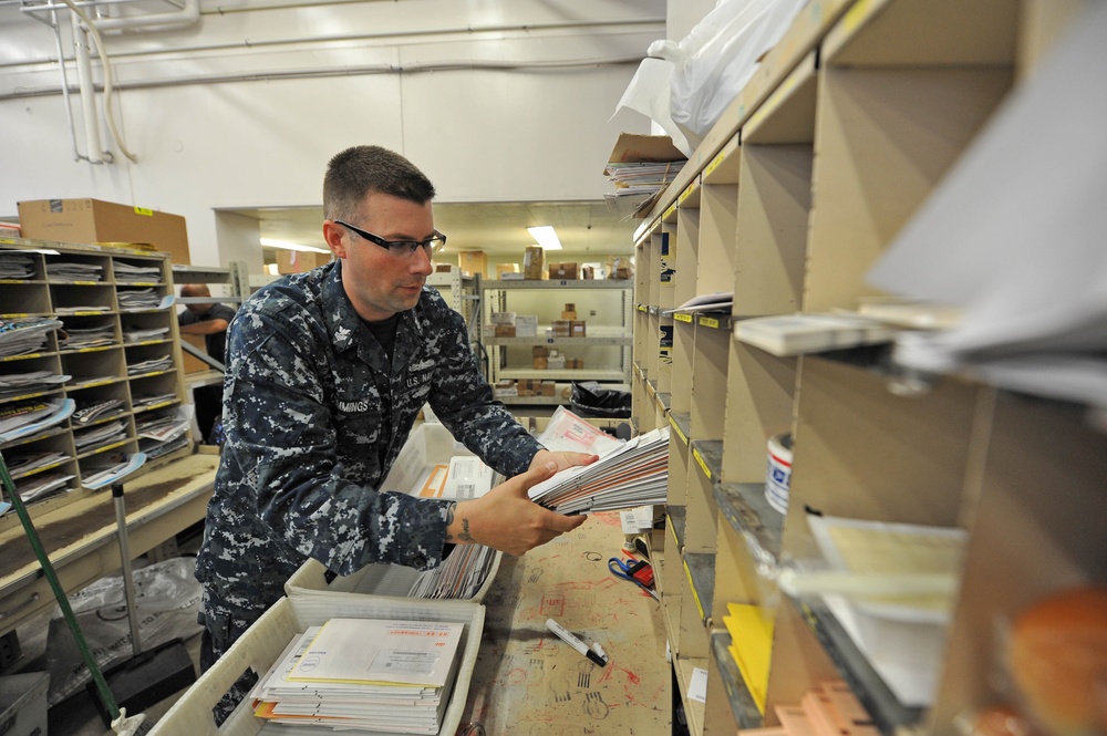 A U.S. Navy sailor in digital camouflage (NWU) uniform is sorting mail in what appears to be a military postal facility. The sailor is handling a stack of envelopes and letters, placing them into or retrieving them from shelving units. The workspace is filled with mail sorting equipment, shelves, and various packages typical of a military post office operation.