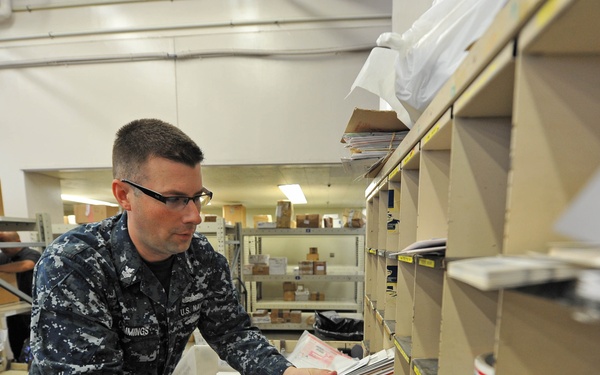 A U.S. Navy sailor in digital camouflage (NWU) uniform is sorting mail in what appears to be a military postal facility. The sailor is handling a stack of envelopes and letters, placing them into or retrieving them from shelving units. The workspace is filled with mail sorting equipment, shelves, and various packages typical of a military post office operation.