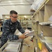 A U.S. Navy sailor in digital camouflage (NWU) uniform is sorting mail in what appears to be a military postal facility. The sailor is handling a stack of envelopes and letters, placing them into or retrieving them from shelving units. The workspace is filled with mail sorting equipment, shelves, and various packages typical of a military post office operation.