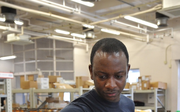 A man in a dark blue t-shirt is sorting mail and packages at what appears to be a military or postal mail handling facility. He is holding orange 'Thriftbooks' padded mailers in one hand while sorting through a pile of packages that includes USPS Priority Mail envelopes. The background shows shelving units stocked with boxes and additional workers in the facility.