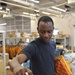 A man in a dark blue t-shirt is sorting mail and packages at what appears to be a military or postal mail handling facility. He is holding orange 'Thriftbooks' padded mailers in one hand while sorting through a pile of packages that includes USPS Priority Mail envelopes. The background shows shelving units stocked with boxes and additional workers in the facility.