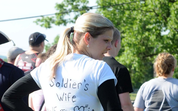 A blonde woman stands at what appears to be an outdoor community event or fun run, wearing a white t-shirt with the handwritten message 'Soldier's Wife Half my ❤️ is Deployed' on the back. She is pushing a pink stroller containing a baby doll. Several other participants are visible in the background.