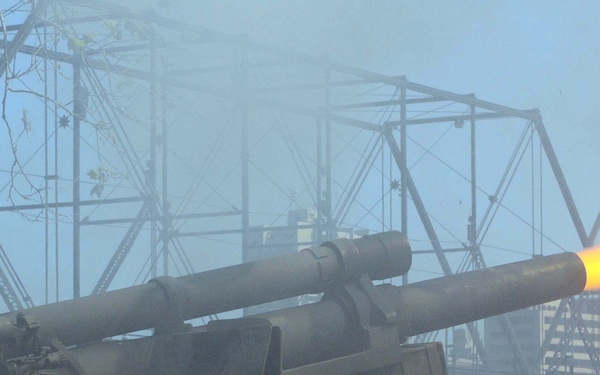 U.S. Army soldiers are firing a ceremonial artillery cannon, with a visible muzzle blast and flame extending from the barrel. The scene appears to take place near a steel truss bridge over a river, suggesting a formal military ceremony or salute. Smoke and fire from the discharge are clearly visible, indicating the cannon was just fired.