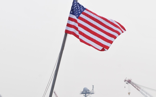 A naval harbor scene showing two personnel in blue uniforms and white hard hats standing on a smaller vessel flying a large American flag. In the background, a large aircraft carrier bearing the hull number '73' is moored at the pier. Industrial cranes and green hillside vegetation are visible in the background, suggesting a naval base in a coastal, possibly East Asian, location.