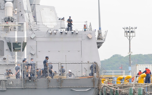The image shows the USS McCampbell, a U.S. Navy Arleigh Burke-class guided-missile destroyer, docking at a pier. Navy personnel are visible on the ship's deck engaged in mooring operations, while dock workers in safety gear assist from the pier. Large black rubber fenders are positioned between the ship's hull and the dock to prevent damage during berthing.